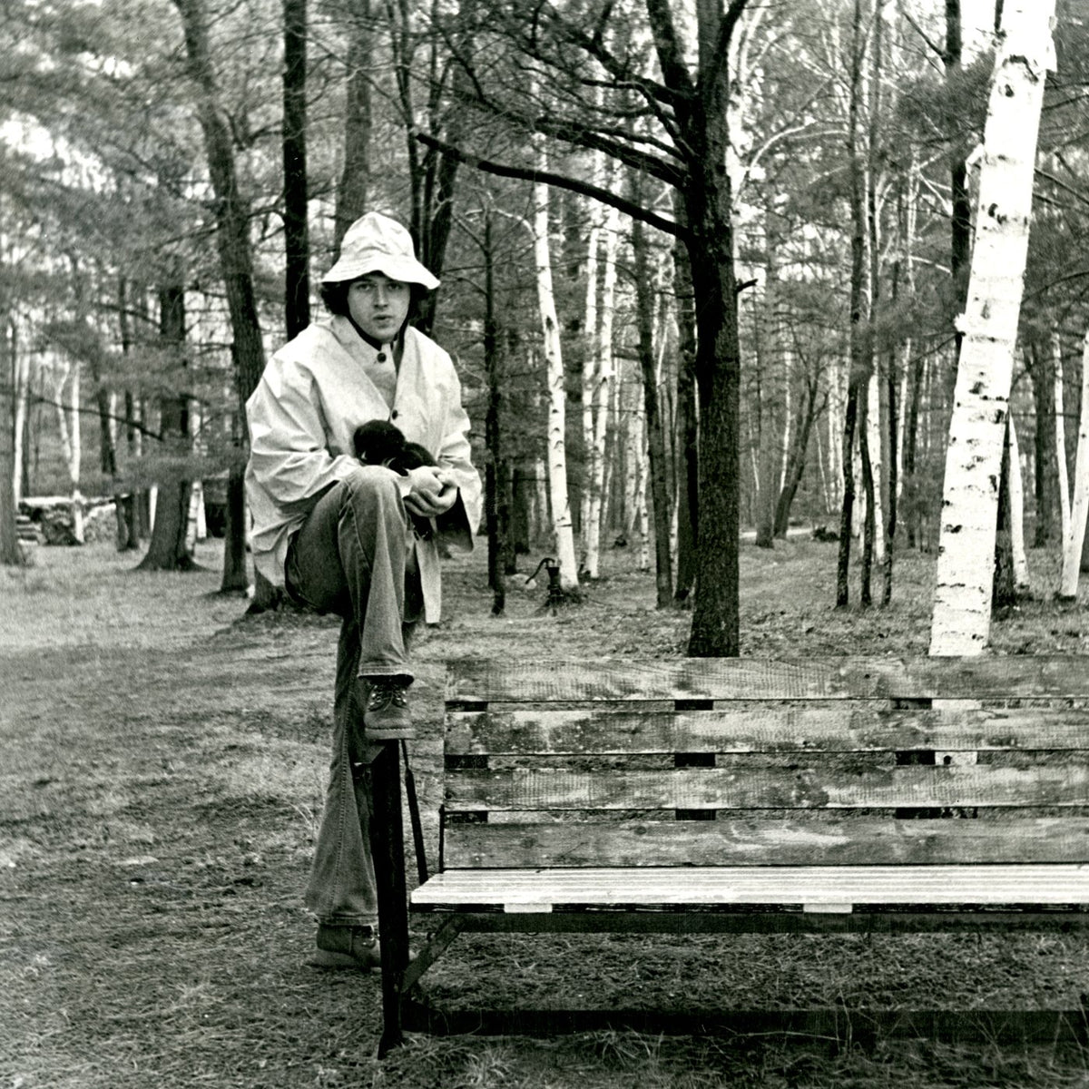 Black and white photo of a man in a raincoat standing by a wooden bench in a forest, Lucky Strikes and Liquid Gold